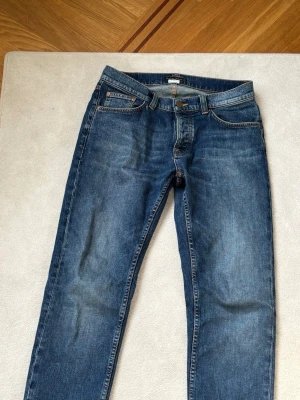 Solid Ollie Wild Blue från Nudie - Säljer ett par klassiska blå jeans från Nudie Jeans med fem fickor och snygga kontrastsömmar. Jeansen har en rak passform och är tillverkade i denim med lätt tvättad look. Bakfickorna har Nudies ikoniska broderi.