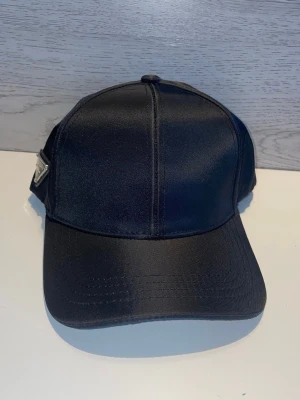 Svart keps från Prada - Snygg svart keps från Prada med klassisk böjd skärm och diskret logga på sidan. Kepsen är tillverkad i ett glansigt tyg som ger en lyxig känsla. Justerbar baktill för perfekt passform. Perfekt accessoar för en trendig streetstyle-look.