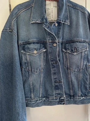 Blå jeansjacka från Acne Studios - Klassisk blå jeansjacka från Acne Studios med raka linjer, två bröstfickor med lock och silverfärgade knappar. Jackan har en tidlös design och är tillverkad i 100% bomullsjeans. Perfekt för dig som gillar en stilren och avslappnad look.
