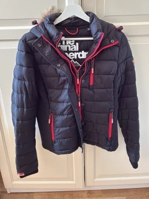 Svart pufferjacka från Superdry - Snygg svart pufferjacka från Superdry med röda dragkedjor och detaljer. Jackan har huva med fuskpäls, flera fickor och quiltad design. Den har en sportig look med logga på ärmen. Storlek L, men passar som M. Mycket fint skick. 