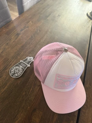 Chrome hearts - Säljer en snygg truckerkeps från Chrome Hearts i rosa och vitt. Kepsen har ett broderat märke framtill med logga och text, samt mesh-tyg baktill för extra ventilation. Justerbar snapback och cool metallknopp på toppen. Perfekt för dig som vill sticka ut. Priset kan diskuteras vid snabb affär😁