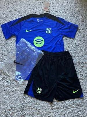 Blå FC Barcelona träningsset Nike [FAST PRIS] - Säljer ett blått FC Barcelona träningsset från Nike med matchande t-shirt och shorts. Tröjan har klubbmärke, Nike-logga och Spotify-logga i neongult samt svarta detaljer på axlarna. Shortsen är svarta med klubbmärke och Nike-logga. Perfekt för fotbollsträning!