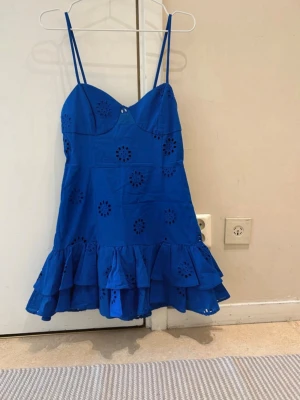  Ny slutsåld Blå kort klänning med volang - Snygg blå kort klänning med tunna axelband och volang nedtill. Klänningen har broderade blommönster och är tillverkad i bomull, vilket gör den perfekt för varma dagar. Söt och trendig modell som ger en somrig vibe.