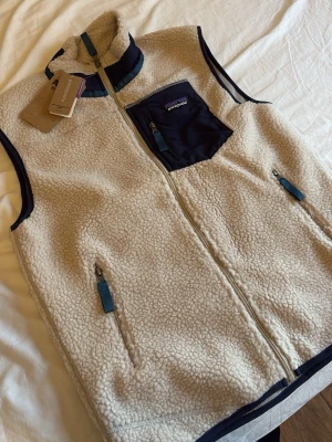Beige fleeceväst från Patagonia - Snygg beige fleeceväst från Patagonia med marinblå detaljer och dragkedjor. Västen har en hög krage, två sidofickor med dragkedja och en bröstficka i kontrastfärg. Perfekt för lager-på-lager och outdoor vibes.