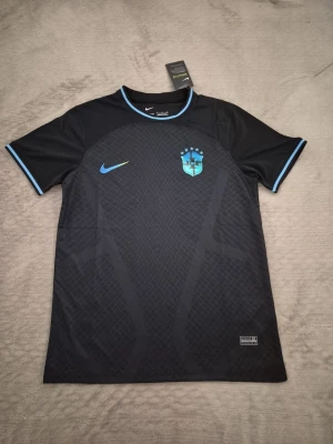 Svart Brasilien Nike fotbollströja - Snygg svart fotbollströja från Nike med Brasilien-emblem och ljusblå detaljer på krage, ärmslut och logga. Tillverkad i lätt och ventilerande material, perfekt för träning eller match. Modern passform med korta ärmar och diskret mönster på framsidan. !Kan passa M!