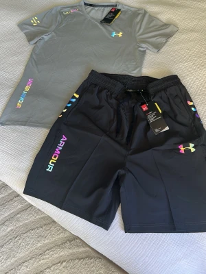 Under Armour träningsset t-shirt & shorts - Snyggt träningsset från Under Armour med en grå t-shirt och svarta shorts. T-shirten har tryck i flera färger och logga på bröstet, medan shortsen har elastisk midja och färgglada detaljer längs sidorna. Perfekt för gym eller löpning.