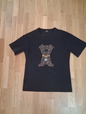 Svart Fendi t-shirt med hundmotiv - Svart t-shirt från Fendi med ett stort broderat hundmotiv på bröstet. Hunden har Fendis klassiska logomönster i brunt och svart samt ett gult halsband med Fendi Roma-tag. T-shirten har rund hals och korta ärmar. Perfekt för dig som gillar statement-plagg.