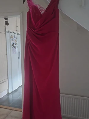 Lång rosa klänning med fjädrar - Snygg lång skrikrosa klänning med draperad framsida och v-ringning. Ena axeln är dekorerad med fjädrar för en extra cool touch. Perfekt för dig som vill sticka ut och känna dig glamorös. Klänningen har en figurnära passform och är ärmlös. Passar storlek 42 också☺️💕