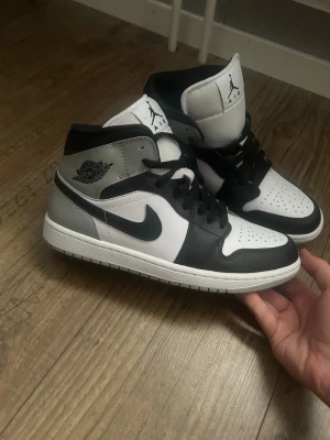 Nike Air Jordan 1 Mid svart/vit/grå - Säljer ett par Nike Air Jordan 1 Mid sneakers i svart, vitt och grått. Skorna har klassisk hög siluett, svart Swoosh, perforerad tåbox och ikoniska Air Jordan-loggan på sidan. Ovandelen är i läder och textil, med svart snörning och vit yttersula.