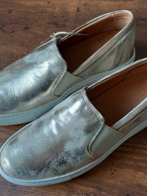 Silvriga loafers med vit sula från Ten Points storlek 36 - Snygga loafers i metallic-silver med vit sula och elastiska partier på sidorna för enkel påtagning. Skorna har en stilren och modern design med rund tå och slät ovandel. Perfekta för dig som vill sticka ut med en glansig touch.