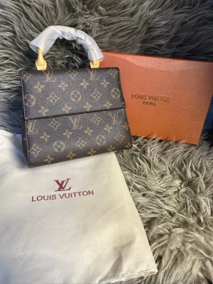  monogram handväska - Snygg handväska i klassiskt brunt monogrammönster med guldiga detaljer och kort handtag. Väskan är tillverkad i tåligt canvasmaterial och har en strukturerad, boxig form. Perfekt för dig som vill ha en ikonisk accessoar med lyxig känsla.