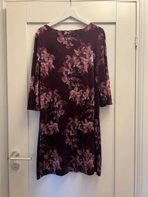 Vinröd blommig klänning med trekvartsärm - Vinröd klänning med stora rosa blommor och trekvartsärm. Rak passform och rund halsringning. Mjukt material som känns skönt mot huden. Perfekt för dig som gillar färg och mönster.