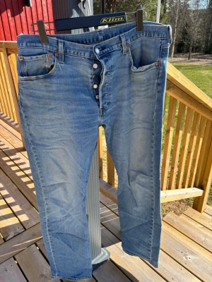 Levi's 501 ljusblå jeans herr - Klassiska Levi's 501 jeans i ljusblå denim med raka ben och knappgylf. Jeansen har fem fickor och är tillverkade i 98% bomull 2% elastan. Perfekta för en avslappnad och tidlös stil.