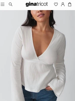 Vit omlottopp från Gina Tricot - Snygg vit omlottopp från Gina Tricot med lång ärm och djup v-ringning. Toppen har knyt i midjan och är tillverkad i ett lätt, strukturerat material som ger en trendig look. Perfekt att styla med jeans eller kjol för en fräsch outfit.