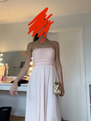 Ljusrosa balklänning - Säljer en elegant ljusrosa axelbandslös långklänning med markerad midja och veckad överdel. Klänningen har en flowy kjol och fina pärldetaljer vid midjan. Perfekt för speciella tillfällen och bal.