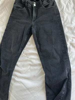 Svarta raka jeans med mid waist - Snygga svarta jeans med raka ben och mid midja. Jeansen har klassisk femficksdesign, dubbla knappar i midjan och subtila slitningar. Perfekta för en avslappnad och trendig look.