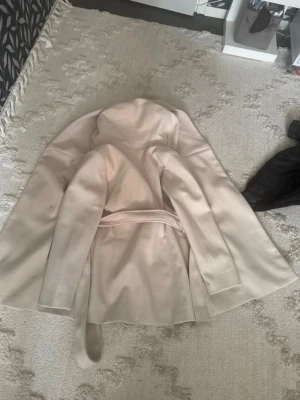 Beige kappa med huva och knytskärp - Stilren beige kappa med huva och knytskärp i midjan. Kappan har knappar på sidan och är tillverkad i ett mjukt tyg som ger en snygg siluett. Perfekt för dig som gillar en enkel men trendig look. Passar i Xs och S