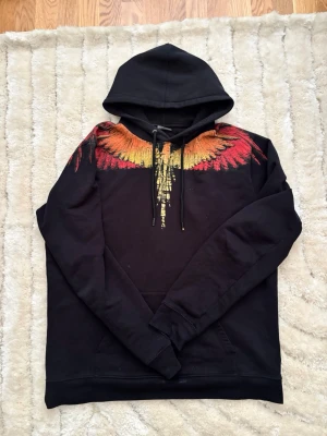 Marcelo burlon hoodie - Svart hoodie med stor huva och känguruficka. Snyggt tryck av vingar i rött, orange och gult över axlar och bröst, som fortsätter på ryggen. Perfekt för dig som vill sticka ut med en unik och färgstark look.