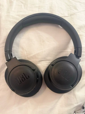 JBL tune 760NC - Snygga svarta over-ear hörlurar från JBL med vadderad bygel och mjuka öronkuddar i konstläder. Perfekta för att lyssna på musik eller poddar med bra ljudkvalitet. Trådlös design och stilren look som passar till allt.