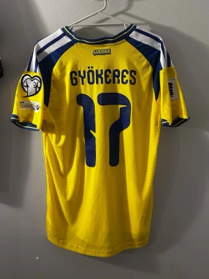 Svensk landslagströja Gyökeres #17 - Gul och blå svensk landslagströja från Adidas med Gyökeres och nummer 17 på ryggen. Tröjan har officiella märken, broderad landslagssköld och UEFA Foundation-märke på ärmen. Tillverkad i lätt och ventilerande funktionsmaterial.