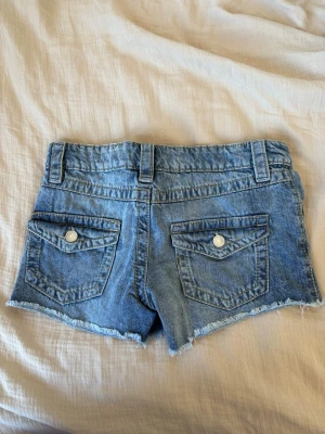 Blå jeansshorts med rå kant från Gina Tricot - Snygga blå jeansshorts från Gina Tricot med rå, fransig kant och klassiska fickor både fram och bak. Shortsen har bälteshällor och knäppning med knapp och dragkedja. Perfekta för varma dagar och en avslappnad stil.