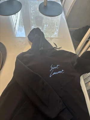 Svart Saint Laurent hoodie - Säljer nu min svarta hoodie från Saint Laurent med blått unikt tryck på bröstet. I princip aldrig använd då den är för liten för mig. Pris går att diskutera vid snabb affär😁 Storlek M