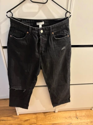 Svarta slitna jeans från H&M - Svarta jeans från H&M i rak modell med slitningar på benet och vid fickan. Klassisk femficksdesign och knappgylf. Jeansen är tillverkade i bomull och har en cool, avslappnad vibe.