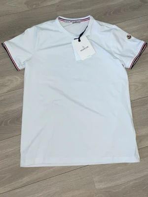Vit Moncler t-shirt med logga - Vit t-shirt från Moncler med klassisk rund hals och korta ärmar. Ärmsluten har snygga ränder i rött och blått samt Moncler-logga på vänster ärm. Tillverkad i mjuk bomull för en skön känsla och stilren look.