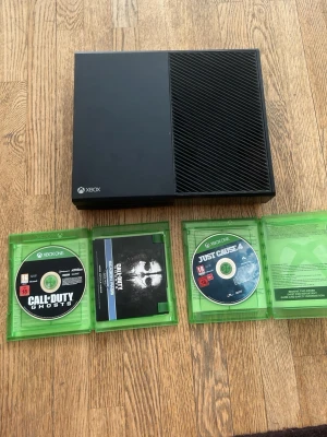 Xbox One konsol med spel - Säljer en svart Xbox One konsol med stilren, rektangulär design i plast. Medföljer spelen Call of Duty: Ghosts och Just Cause 4, båda i sina gröna originalfodral. Perfekt för dig som vill ha en klassisk spelupplevelse med populära titlar.