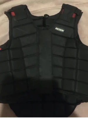 Svart skyddsväst från Jacson - Säljer en svart skyddsväst från Jacson med vadderad och segmenterad design för extra rörlighet. Västen har justerbara axelband och kardborre på sidorna. Perfekt för ridning eller andra aktiviteter där extra skydd behövs. Den är storlek 10år. Lite hår och skit på karborrbanden men tar bort så mycket jag kan. 