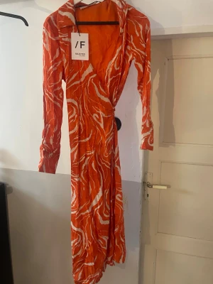 Orange omlottklänning från Selected Femme - Säljer en lång orange omlottklänning från Selected Femme med vitt abstrakt mönster. Klänningen har lång ärm, krage och knyt i midjan för en snygg siluett. Perfekt för dig som vill sticka ut med färg och mönster.