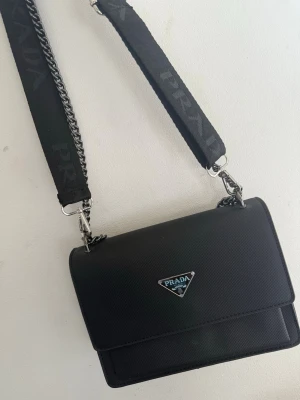Svart axelväska lyxig  - Snygg svart axelväska med klassisk rektangulär form och silverfärgade detaljer. Väskan har en bred axelrem i tyg med kedjedetaljer och ikoniska logga framtill. Tillverkad i slitstarkt material med stilren design.