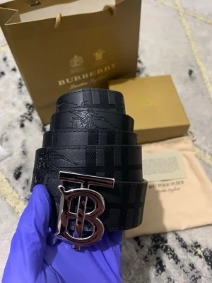 Burberry bälte med TB-spänne -  snygg Burberry-bälte med ikoniskt TB-spänne i metall. En sida är svart med diskret rutmönster och den andra sidan är beige med klassiskt rutig design. Båda sidor är tillverkade i skinn och har exklusiv känsla, perfekta för att lyfta din outfit.