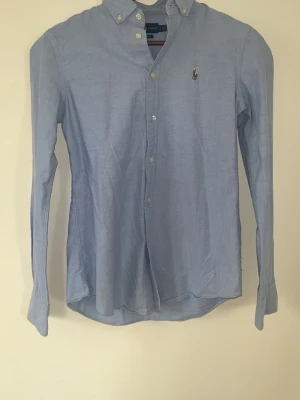 Blå skjorta från Ralph Lauren - Klassisk ljusblå skjorta från Ralph Lauren i slim fit som är perfekt nu inför simmaren. Skick 9/10, otroligt bra skick på denna ! Säljer för att den är för liten för mig. Passar ca 172-177 cm. Hör av dig vid minsta fundering! 