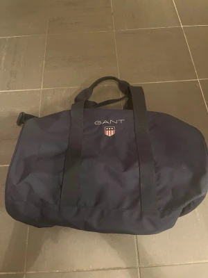 Marinblå weekendbag från GANT - Snygg marinblå weekendbag från GANT med broderad logga och emblem på framsidan. Väskan har två kraftiga handtag, avtagbar axelrem och cylinderformad design. Tillverkad i slitstarkt tygmaterial, perfekt för kortare resor eller gymmet.