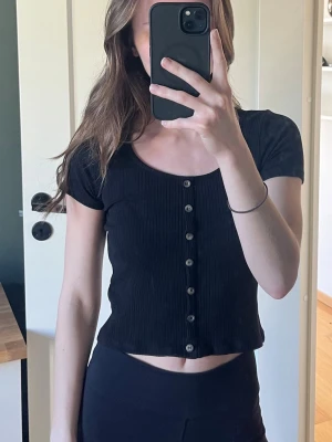 Svart ribbad croppad topp - Snygg svart croppad topp med ribbad struktur och knappar framtill. Kortärmad modell med rund halsringning, perfekt till jeans eller kjol. Enkel och stilren design som passar till många olika looks.