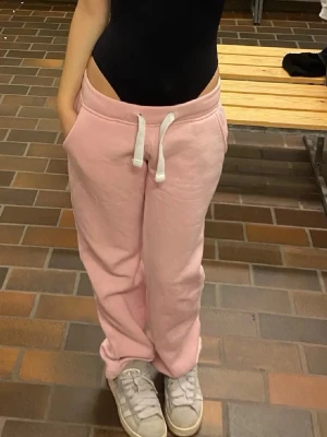 Ljusrosa mjukisbyxor från russemerch - Säljer ett par ljusrosa mjukisbyxor med vita dragsnören i midjan och bekväm passform. Byxorna har resår i midjan, raka ben och är gjorda i mjukt sweatshirtmaterial. Perfekta för chill dagar eller när du vill vara extra bekväm.