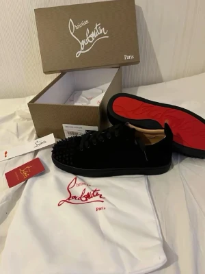 Svarta Christian Louboutin sneakers - Snygga svarta sneakers från Christian Louboutin med klassiska röda sulor och coola nitar på tån. Skorna har en låg profil, svarta skosnören och är tillverkade i mocka med läderdetaljer. Perfekta för dig som vill sticka ut med exklusiv design.