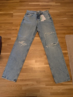 Ljusblå ripped jeans med raka ben - Säljer ett par ljusblå jeans med raka ben och stora slitningar på båda benen. Jeansen har klassisk femficksmodell, hög midja och är tillverkade i denim. Perfekta för en avslappnad och trendig look.
