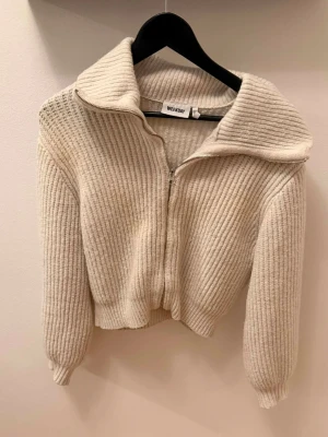 Beige stickad ziptröja från Weekday - Säljer en beige stickad ziptröja från Weekday med bred krage och ribbstickat mönster. Tröjan har långa ärmar, dragkedja framtill och en croppad passform. Perfekt för lager-på-lager och chill stil.