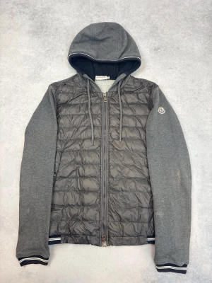 Grå Moncler cardiganjacka med huva - Snygg grå cardiganjacka från Moncler med quiltad front och mjuka ärmar i sweatshirtmaterial. Jackan har huva med dragsko, hel dragkedja och ribbade muddar. Moncler-logga på ärmen ger en exklusiv touch. Perfekt för lager på lager och streetwear.