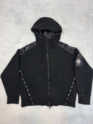 Svart hoodie från Moncler med detaljer - Snygg svart hoodie från Moncler med dragkedja, huva och glänsande nylonpaneler på axlar och huva. Stora fickor framtill och Moncler-logga på ärmen. Dragsko i huvan och detaljerade band längs fickorna. Perfekt för en trendig streetwear-look.