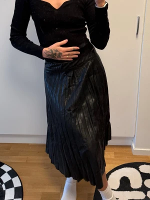 Svart plisserad långkjol i skinnimitation - Säljer en svart plisserad långkjol i skinnimitation. Kjolen har hög midja och ett snyggt fall med markerade veck. Perfekt att styla med en croppad topp eller stickad tröja för en trendig look.