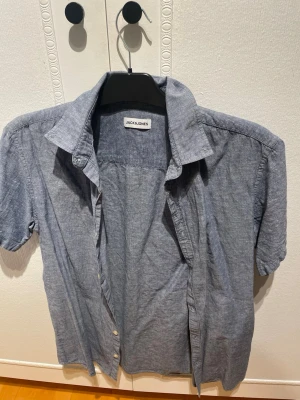 Blå kortärmad skjorta Jack & Jones - Snygg blå skjorta från Jack & Jones i storlek M. Skjortan har korta ärmar, klassisk krage och knappar framtill. Perfekt till sommarens alla tillfällen och lätt att matcha med olika outfits.