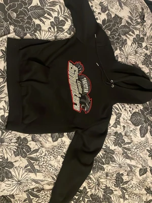 Svart hoodie med tryck från TrapstarSHARKS - Säljer en svart hoodie från TrapstarSHARKS med stort tryck på bröstet i rött, vitt och grått. Hoodien har huva med dragsko och en klassisk känguruficka framtill. Perfekt för dig som gillar streetwear och vill ha en bekväm och snygg tröja.
