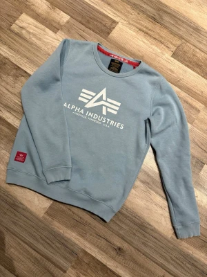 Ljusblå sweatshirt Alpha Industries - Ljusblå sweatshirt från Alpha Industries med vitt tryck på bröstet och rund hals. Inga prisförslag är dumma!
