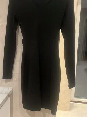 Svart långärmad kortklänning - Stilren svart klänning med lång ärm och kort längd. Klänningen har en omlottdetalj i ryggen som ger en snygg twist. Passformen är kroppsnära och tyget känns mjukt och stretchigt, perfekt för dig som gillar en enkel men elegant look. Uppfattas s i storlek 