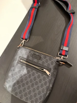 Gucci axelväska med GG-mönster - Snygg svart axelväska från Gucci med klassiskt GG-mönster över hela väskan. Väskan har en justerbar axelrem i blått och rött, samt en praktisk ytterficka med dragkedja. Tillverkad i canvas och läderdetaljer. Perfekt för dig som gillar streetstyle och lyx.