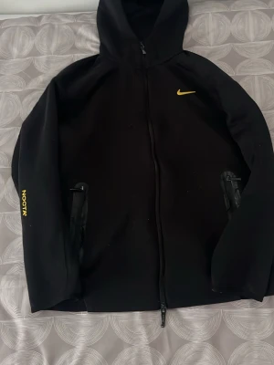 Svart Nike NOCTA hoodie med dragkedja - Säljer en svart hoodie från Nike NOCTA med gul logga på bröstet och NOCTA-tryck på ärmen. Hoodien har hel dragkedja framtill, två fickor med dragkedja och en klassisk huva. Materialet är mjukt och stretchigt, perfekt för en sportig och clean look.