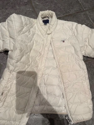 Vit dunjacka från GANT - Snygg vit dunjacka från GANT med quiltat mönster och hög krage. Jackan har dragkedja framtill och liten broderad logga på bröstet. Perfekt för kalla vinterdagar och riktigt stilren look.